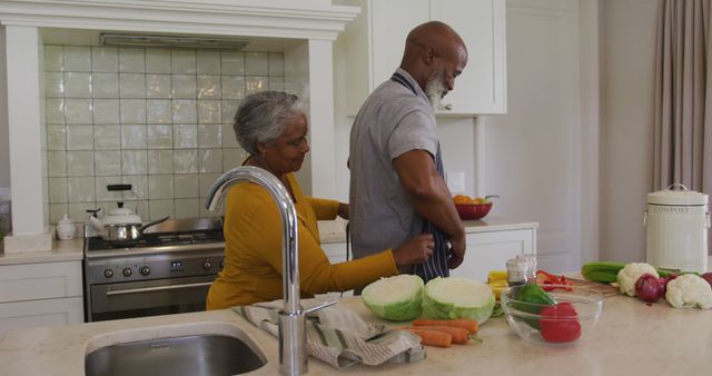 Elderly man and woman preparing vegetables together in a modern kitchen. Both seem engaged, with the woman adjusting the man's apron as they prepare vegetables like cabbage, carrots, and pepper. This setting is ideal for use in content about health, cooking at home, senior lifestyle, elderly activities, and promoting family togetherness.