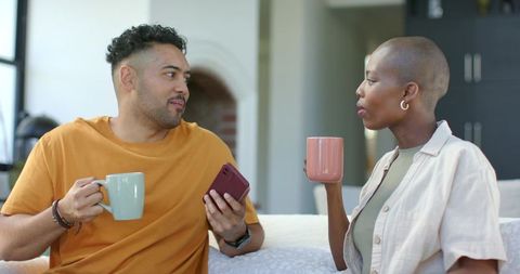 African american couple chatting over coffee on sofa while sharing smartphone and mugs