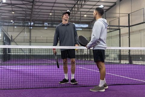 Male players chatting on indoor padel court with purple turf holding paddles and ball