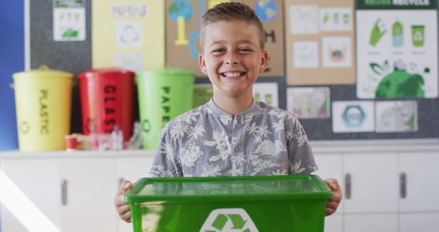 Caucasian schoolboy smiling, holding recycling bin, standing in classroom. children at primary school in summer.