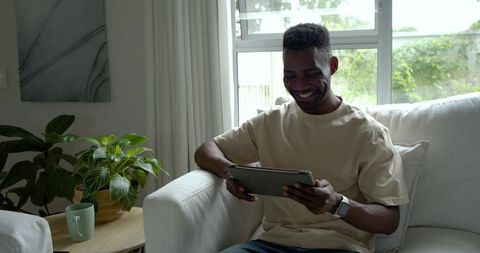 Smiling man sitting on sofa using tablet and checking smartwatch in bright living room