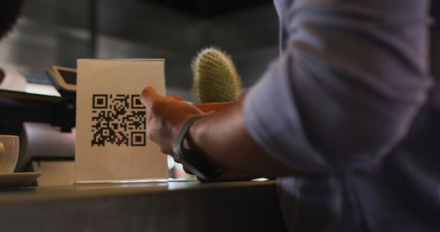 This image shows a person scanning a QR code presented on a stand at a cafe counter, illustrating the use of modern technology for digital payments. Ideal for use in articles, blogs, or promotional materials on modern technology, digital payments, contactless transactions, and tech-savvy lifestyle.
