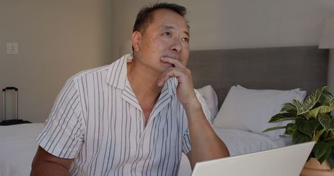 Focused asian man thinking while using laptop in bedroom workspace