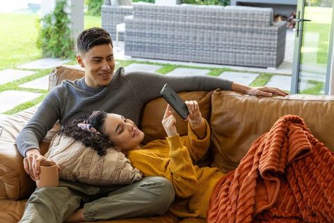 Young couple relaxing on leather sofa, woman scrolling smartphone, cozy sunlit living room