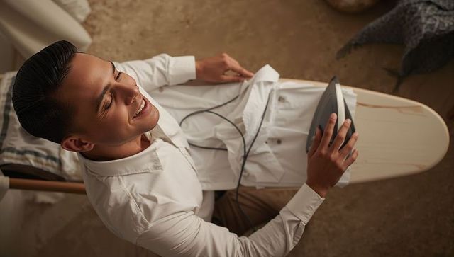 Smiling man ironing dress shirt on board preparing outfit for work morning routine at home