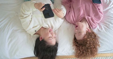 Diverse friends lying on bed laughing and scrolling phones sharing cozy at-home moment