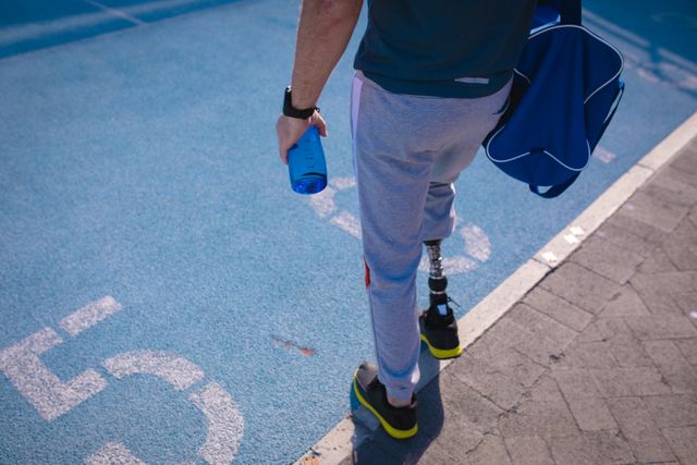 Low section of caucasian disabled male athlete with prosthetic leg holding bottle and bag. professional runner training at sports stadium.