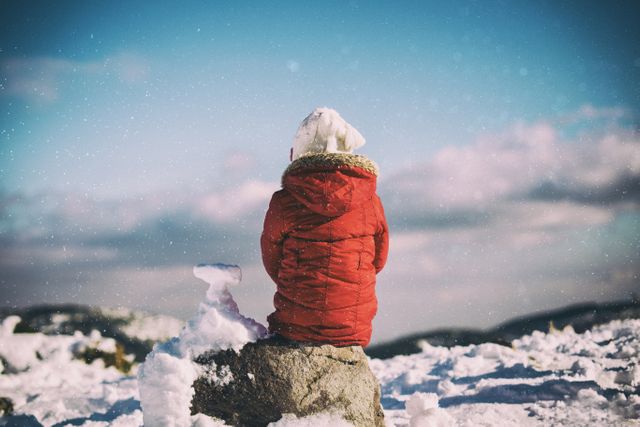 Child dressed in warm red jacket and white hat sits on snow-covered ground, facing snowy mountainous scenery. Ideal for concepts of childhood, winter activities, exploring nature, and seasonal adventures. Perfect for winter clothing advertisements, holiday greetings, and family-oriented content.