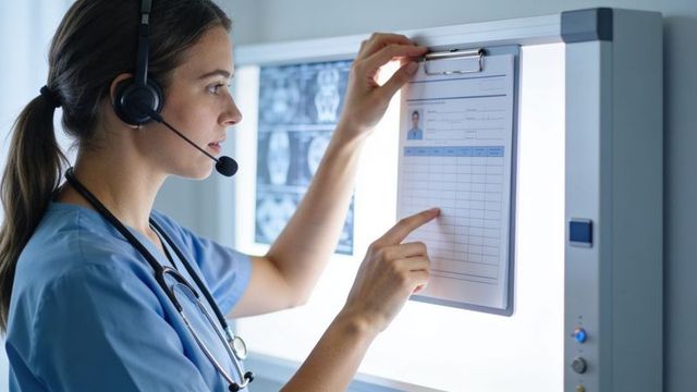 Nurse in blue scrubs wearing headset pointing at patient form on radiology lightbox with stethoscope