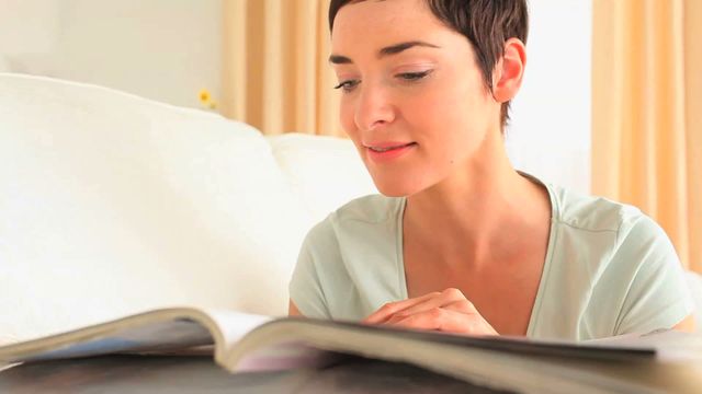 This video captures a brunette woman lying on a sofa reading a magazine in a relaxed home environment. It could be used for editorial content, lifestyle blogs, articles on relaxation, home living, or leisure activities. It is excellent for conveying a sense of calm and comfort in various print and digital media.