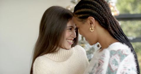 Smiling diverse female couple leaning foreheads together by decorated christmas tree