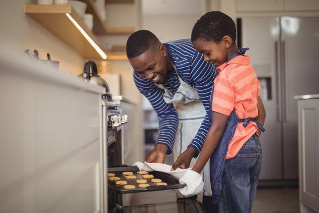 This image captures a joyful moment of a father and son baking cookies together in a modern kitchen. The father is taking a tray of fresh cookies out of the oven while the son assists. Ideal for use in family-oriented advertisements, parenting blogs, cooking websites, and home lifestyle magazines.
