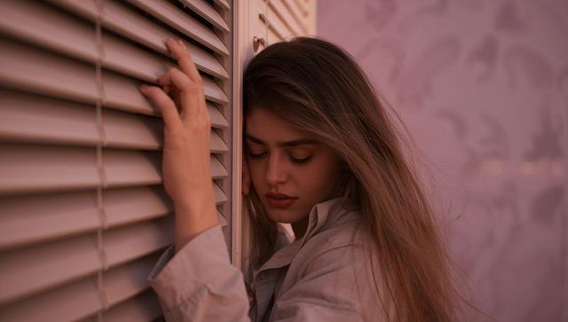Young woman leaning against blinds in moody pink glow, contemplative interior portrait