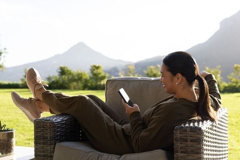 Relaxing woman scrolling smartphone on wicker lounge chair overlooking mountain lawn