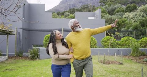 Senior couple walking through landscaped yard, man pointing toward mountain view, smiling