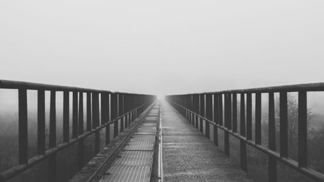 Dramatic perspective showing a bridge extending into thick fog, creating a mysterious and eerie atmosphere. Black and white tones enhance the somber and atmospheric mood. Suitable for themes of mystery, solitude, and journeys into the unknown. Great for use in artistic projects, book covers, promotional materials for thrillers or moody film scenes, and travel or adventure platforms to evoke a sense of intrigue.