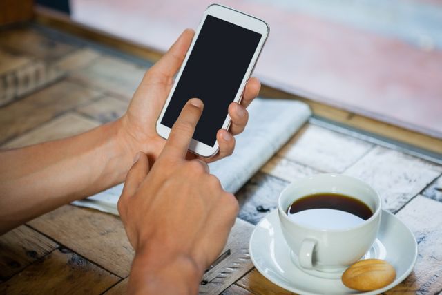 This image shows a woman using a smartphone while sitting at a coffee shop table. Her hand is interacting with the touchscreen, and there is a cup of coffee and a biscuit on the table. This image can be used for themes related to technology, communication, morning routines, relaxation, and lifestyle. It is suitable for websites, blogs, and advertisements focusing on coffee culture, mobile technology, and casual dining.