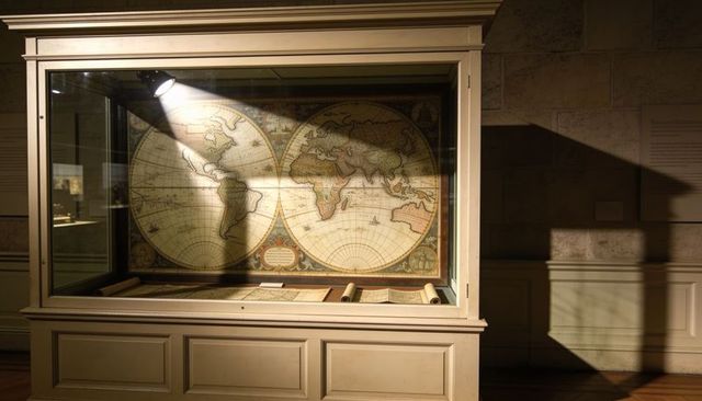 Displaying antique world map and rolled charts in museum glass case under spotlight