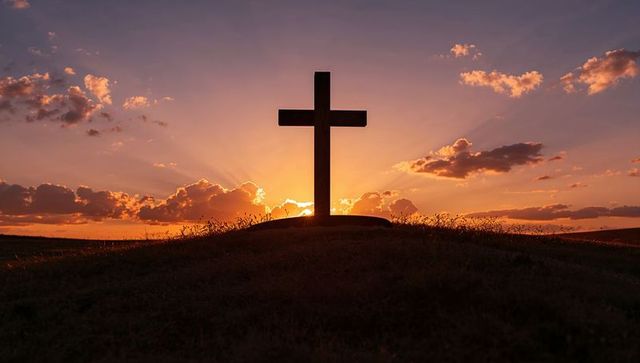 Standing cross silhouette with radiant sunset halo over hilltop and glowing sky