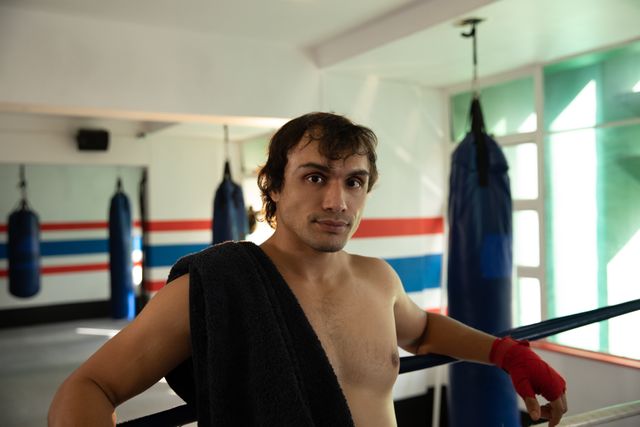 Front view of a young biracial male boxer with short dark hair, shirtless, with towel over his shoulder, standing in a boxing gym, looking straight to the camera.