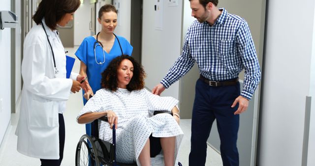 Female patient sitting in wheelchair receiving assistance from medical staff and family member in hospital corridor. Suitable for depicting healthcare scenarios, patient recovery, medical assistance, and rehabilitation.