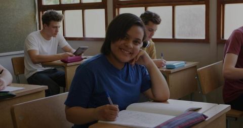 Smiling teen student writing in notebook at classroom desk with tablet and classmates