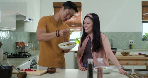 Asian couple preparing fresh salad together in modern kitchen, man sprinkling dressing