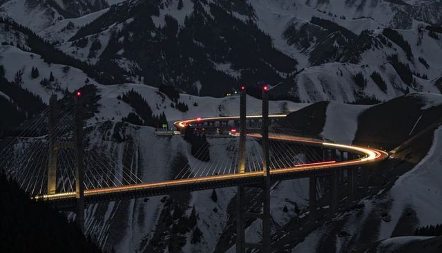 Curving cable-stayed viaduct glowing with vehicle light trails across snowy alpine canyon