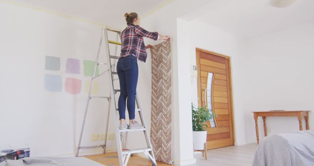 Young woman hanging wallpaper on a white wall using a ladder. Various paint swatches are visible on the wall nearby. This image can be useful for articles or content about home improvement, DIY projects, interior design, and modern living spaces.