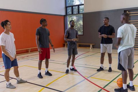 Diverse male teammates talking before pickup basketball game in sunlit indoor gymnasium
