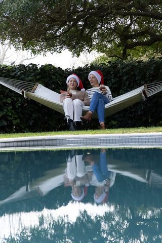 Friends relaxing on backyard hammock by pool wearing santa hats and toasting holiday drinks