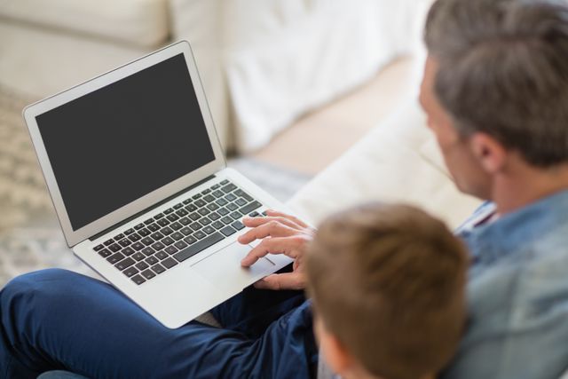 Father and son using laptop in living room, depicting family bonding and use of technology at home. Suitable for themes related to parenting, family life, home education, and digital communication.