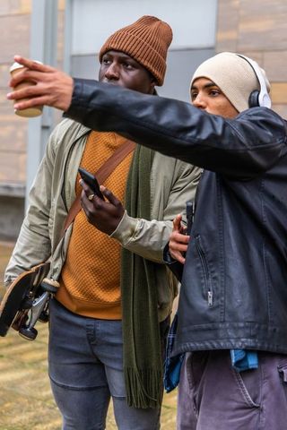 Diverse young men checking smartphone and pointing while holding skateboard and coffee