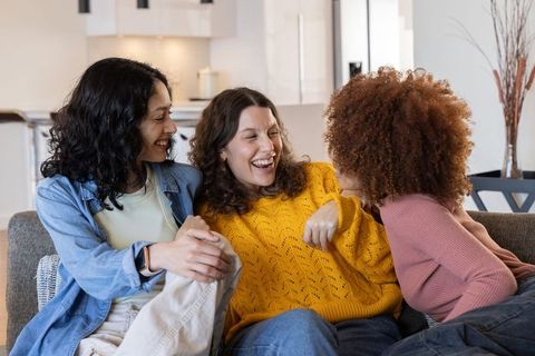 Diverse women laughing and chatting on couch sharing smartwatch and cozy friendship moments