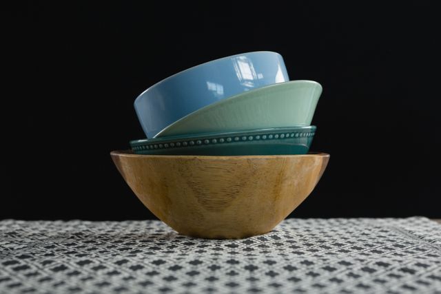 Stacked ceramic bowls in various colors placed on a wooden bowl, set on a patterned tablecloth. Ideal for use in articles or advertisements related to kitchenware, home decor, dining, minimalistic design, or table setting ideas.