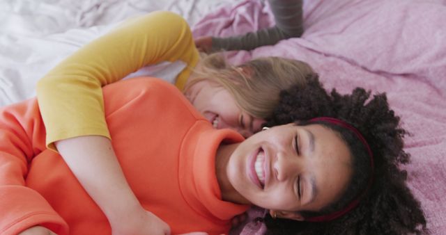 Two friends are lying on a bed, hugging and laughing. Both appear to be teenagers in casual, colorful clothing. This cheerful moment captures the essence of friendship and joy. Ideal for use in lifestyle blogs, advertisements for sleepwear or bedding, and articles about friendship and family.