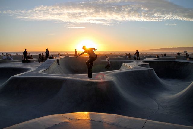 Silhouette of a skateboarder performing a trick against a scenic sunset backdrop at an urban skate park filled with concrete bowls. Ideal for advertising urban culture, youth activities, action sports, and outdoor adventure themes.