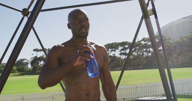 Fit shirtless african american man exercising on climbing frame outdoors, resting and drinking water. cross training for fitness at a sports field.