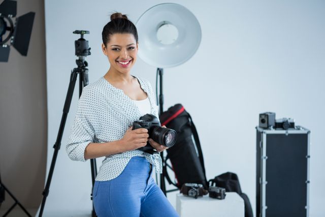 This image shows a young female photographer smiling while holding a digital camera in a professional photography studio. The background includes various photography equipment such as lighting, tripods, and camera gear. This image can be used for articles, blogs, or advertisements related to photography, creative industries, professional photographers, and studio setups.