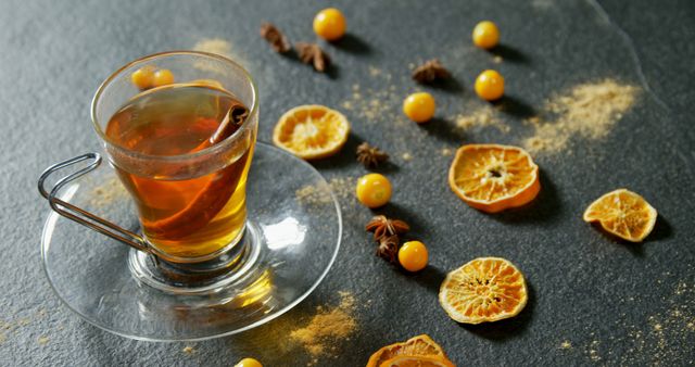 A glass cup of tea with a cinnamon stick is accompanied by dried orange slices and spices on a dark textured surface, with copy space. Warm tones and the arrangement suggest a cozy, aromatic experience ideal for relaxation or cold weather.