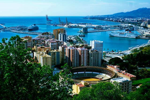 Scenic view showing coastal city with prominent bullring, modern port with large ships, and surrounding urban high-rise buildings against a mountainous backdrop. Useful for travel websites, urban development studies, tourist guides, and real estate promotions highlighting coastal cities.