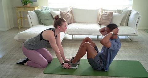 Trainer holding feet while partner doing sit-ups on mat in bright modern living room