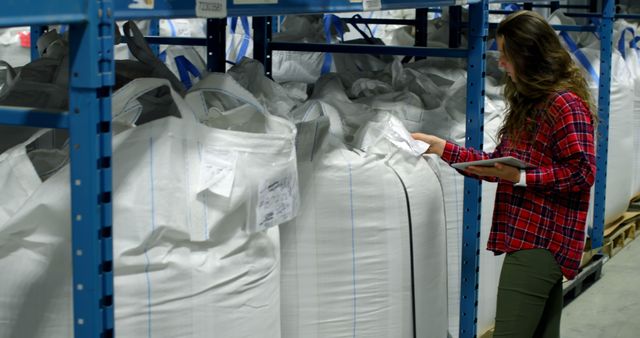 Female warehouse worker in a red plaid shirt holding a tablet while checking large bags stored on shelves. Suitable for use in articles and advertisements related to logistics, supply chain management, warehouse inventory management, and industrial storage solutions.