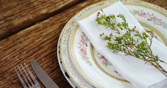 A vintage floral plate is set on a wooden table with a neatly folded napkin adorned with fresh greenery, accompanied by a silver fork. The arrangement suggests a rustic and elegant dining experience, with attention to detail in table setting.