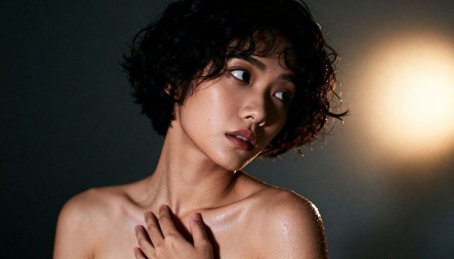 Gazing bare-shouldered asian woman touching chest, dewy skin glowing under warm rim light