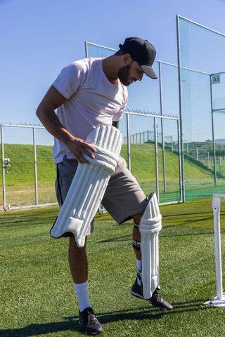 Cricketer fastening batting pads on artificial turf near white stumps and practice nets