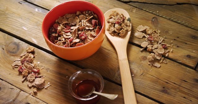 Image shows healthy breakfast with cereal flakes and dried strawberries in orange bowl on rustic wooden table. Wooden spoon is placed beside bowl, with some cereal flakes spread around. Small glass cup with honey and spoon complement breakfast setup. Ideal for healthy eating concept, breakfast, diet promotion, and food blogs.