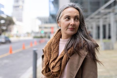 Mature woman standing on urban sidewalk wearing brown coat and knit scarf looking away