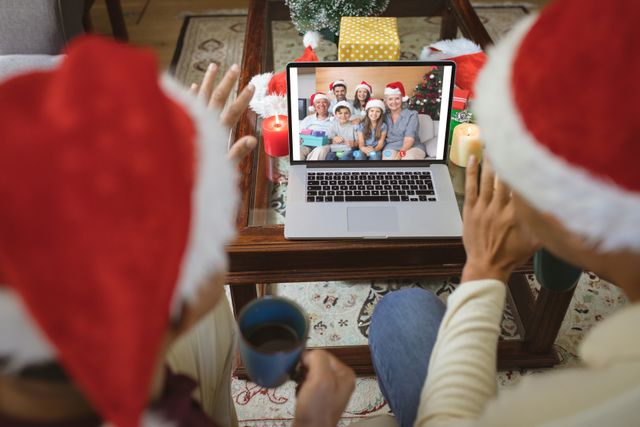 Diverse couple wearing Santa hats talking to family via video call during the Christmas season, using a laptop at home, drinks and Christmas decorations surrounding them. Perfect for marketing campaigns about virtual celebrations, online communication, remote family interactions during holidays, and festive greetings in the digital age.