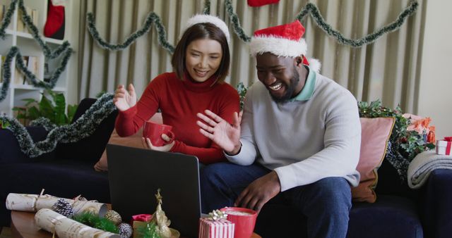 This depicts a couple video chatting on a laptop during a Christmas celebration at home. Both are wearing Santa hats and are surrounded by festive decorations, including garlands and presents. This is perfect for materials related to virtual holiday gatherings, festive celebrations, long-distance communication, and multicultural bonding.